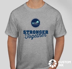 A grey t shirt with the SANYS logo- a circle with a picture of new your state in it and SANYS people with developmental disabilities speaking up for ourselves and others. Underneath it in larger print it say Stronger Together